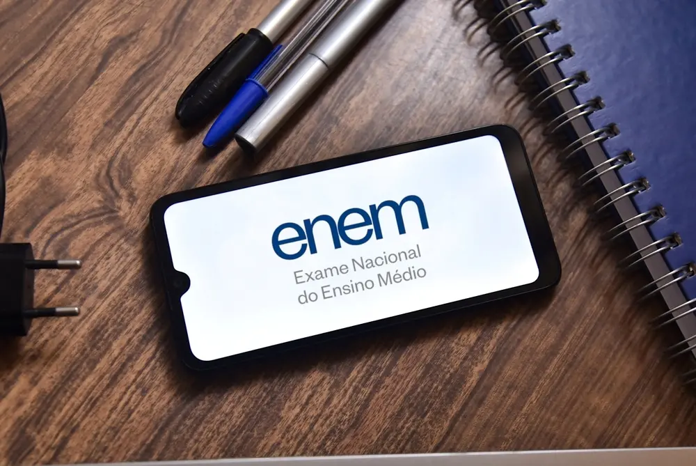 Caderno aberto e canetas sobre uma mesa de madeira, com cartão de identificação mostrando o texto “enem Exame Nacional do Ensino Médio”.