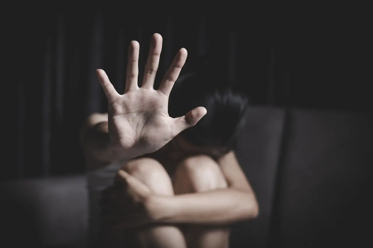Mão de mulher branca fazendo o sinal de pare, com fundo escuro e rosto escondido nos joelhos.