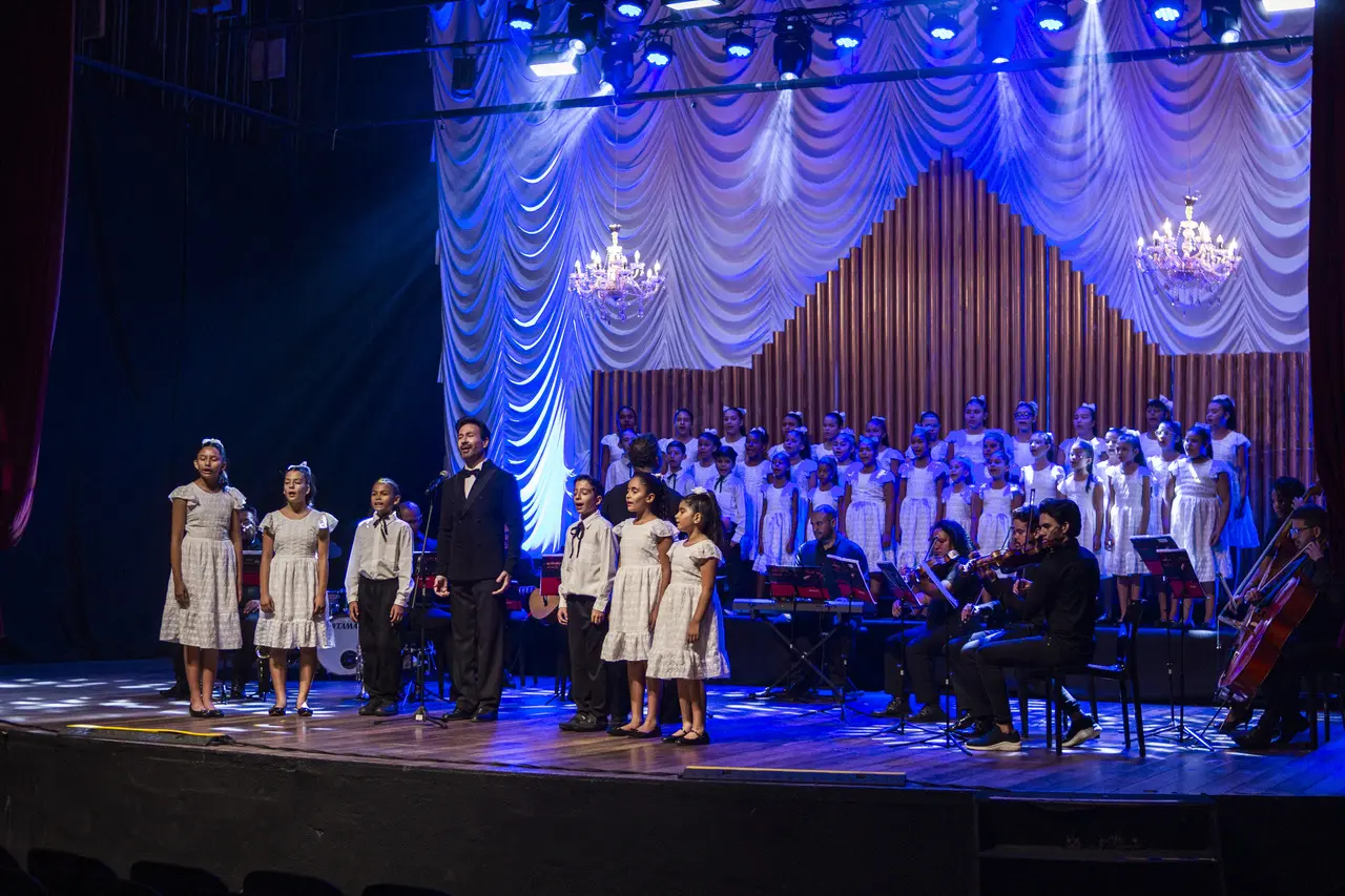 Um grande coro infantil e músicos de uma orquestra se apresentam no palco iluminado de um teatro sob uma forte luz azul e branca. Vestidas de branco, as crianças se dividem entre a frente do palco e uma grande estrutura ao fundo, acompanhadas por um cantor solista em traje formal preto.