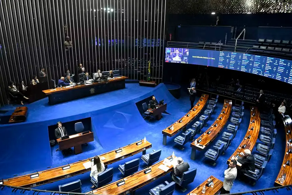 Mulher de blazer escuro e óculos de grau em sessão no plenário, fazendo uso da palavra e segurando documentos, com microfone e mesa ao lado, enquanto ao fundo outros parlamentares trabalham.