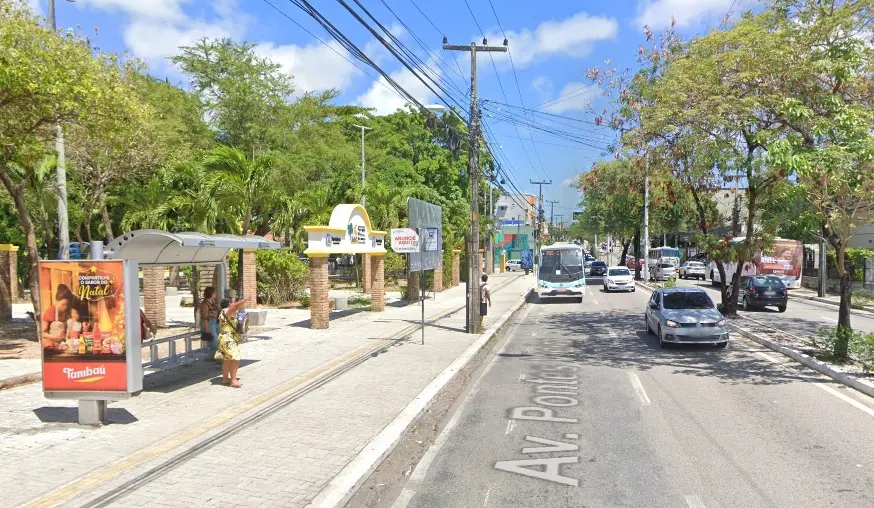 Pedestres aguardam em uma parada de ônibus na calçada de uma avenida movimentada, destacando uma mulher que acena para um ônibus branco que se aproxima ao fundo. O cenário urbano arborizado conta com carros em movimento e a entrada de um parque sob o céu azul, compondo uma cena cotidiana e clara em um dia ensolarado.
