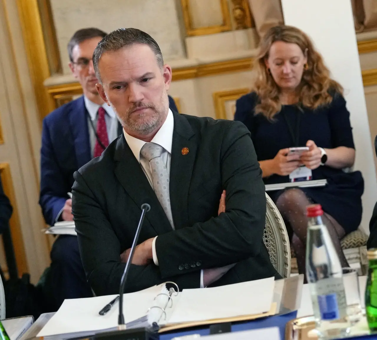 Autoridades participam de uma reunião formal em uma sala elegante, destacando Jamieson Greer, um homem de terno escuro com os braços cruzados e expressão séria em primeiro plano. Outros profissionais sentados ao fundo observam e utilizam aparelhos celulares, compondo uma cena corporativa e focada em um ambiente institucional detalhado.