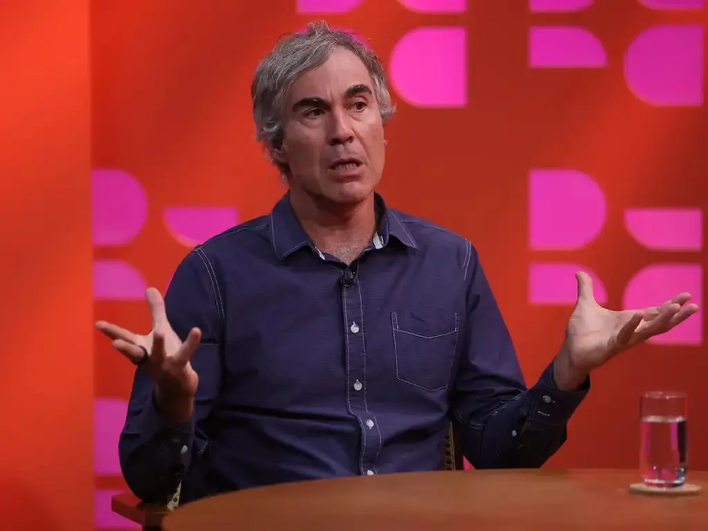 Bruno Paes Manso participa de entrevista em estúdio, sentado à mesa e gesticulando com as mãos enquanto fala. Ao fundo, há um painel em tons de vermelho e rosa.