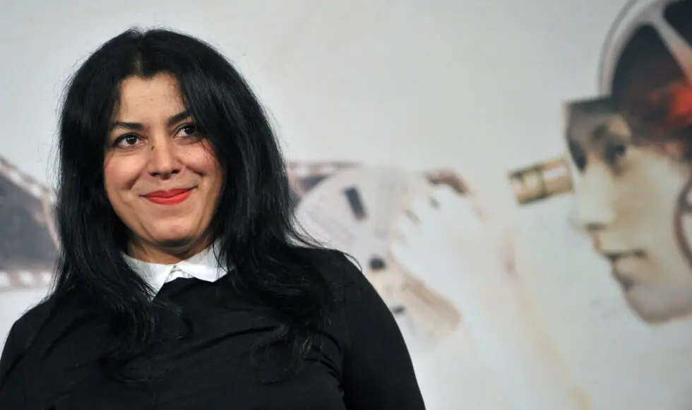 Marjane Satrapi sorri para a câmera durante evento público. A escritora e cineasta aparece em primeiro plano, vestindo roupa preta com gola branca, diante de um painel que exibe a ilustração de um rosto observando por um microscópio.