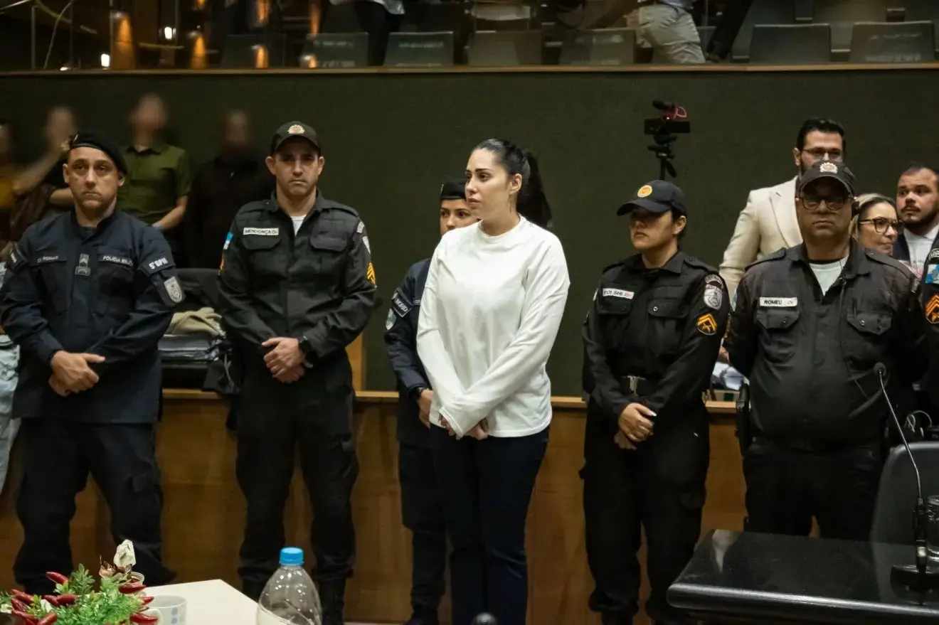 Monique Medeiros em pé no Tribunal do Júri do Rio de Janeiro, vestindo blusa branca e calça escura, cercada por policiais militares durante o julgamento do caso Henry Borel.