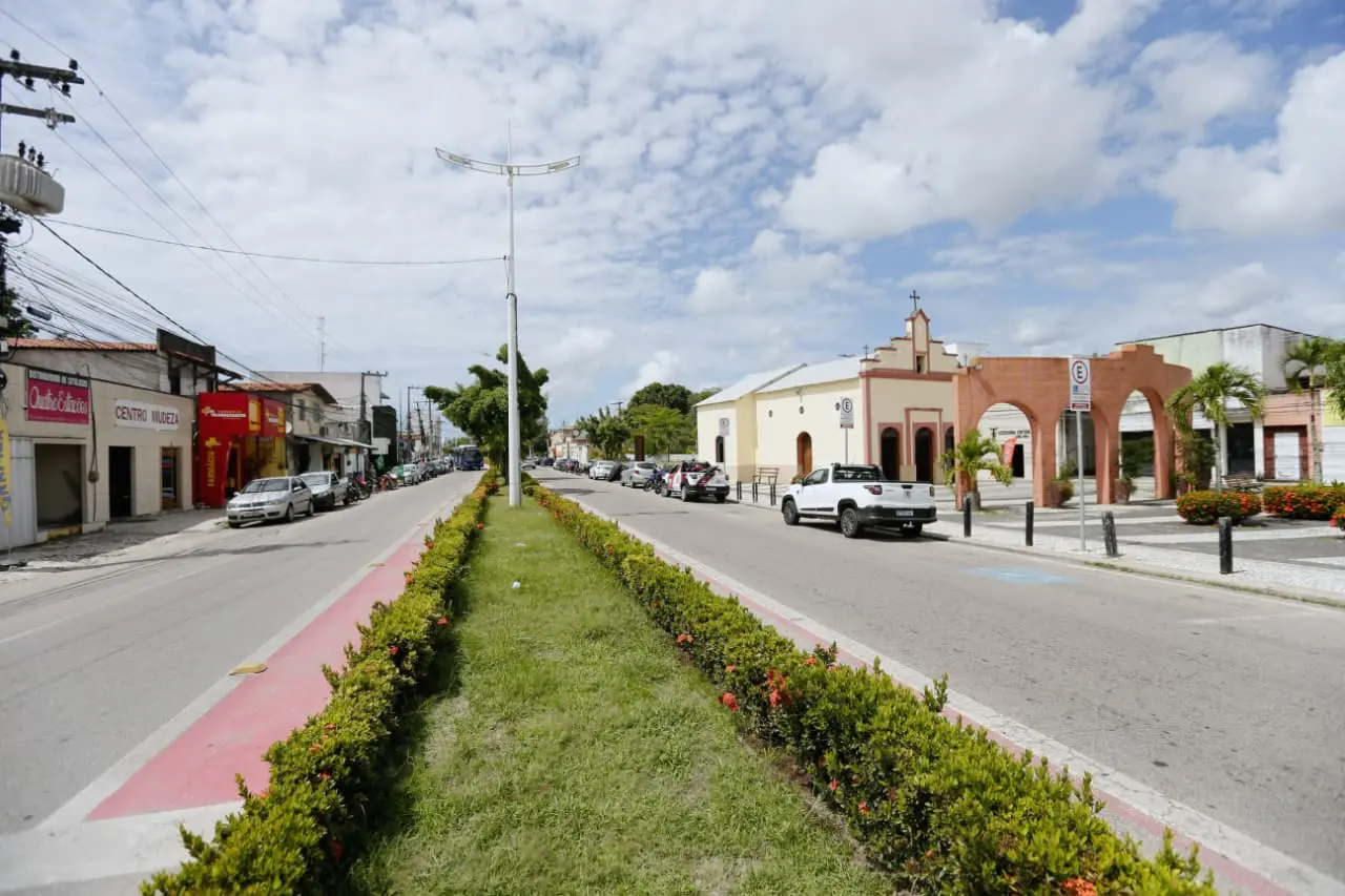 Avenida Eusébio de Queiroz com canteiro central arborizado e ciclovia. À direita, destacam-se uma igreja antiga e uma praça com arcos de tijolos sob céu parcialmente nublado.
