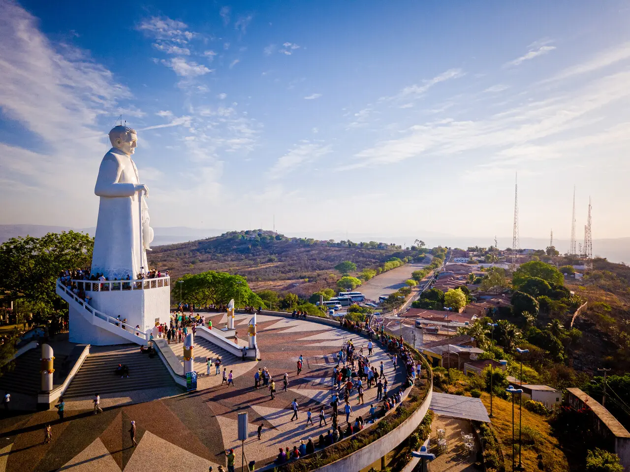 Vista aérea da estátua de Padre Cícero no Horto, em Juazeiro do Norte (CE), durante o dia. O monumento branco se destaca sobre uma grande praça com visitantes circulando e observando a paisagem do entorno. Ao fundo, aparecem áreas urbanas, vegetação e colinas sob céu azul com nuvens dispersas.