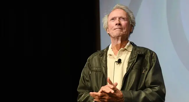 Clint Eastwood aparece em pé sobre um palco, usando jaqueta de couro escura e camisa clara.