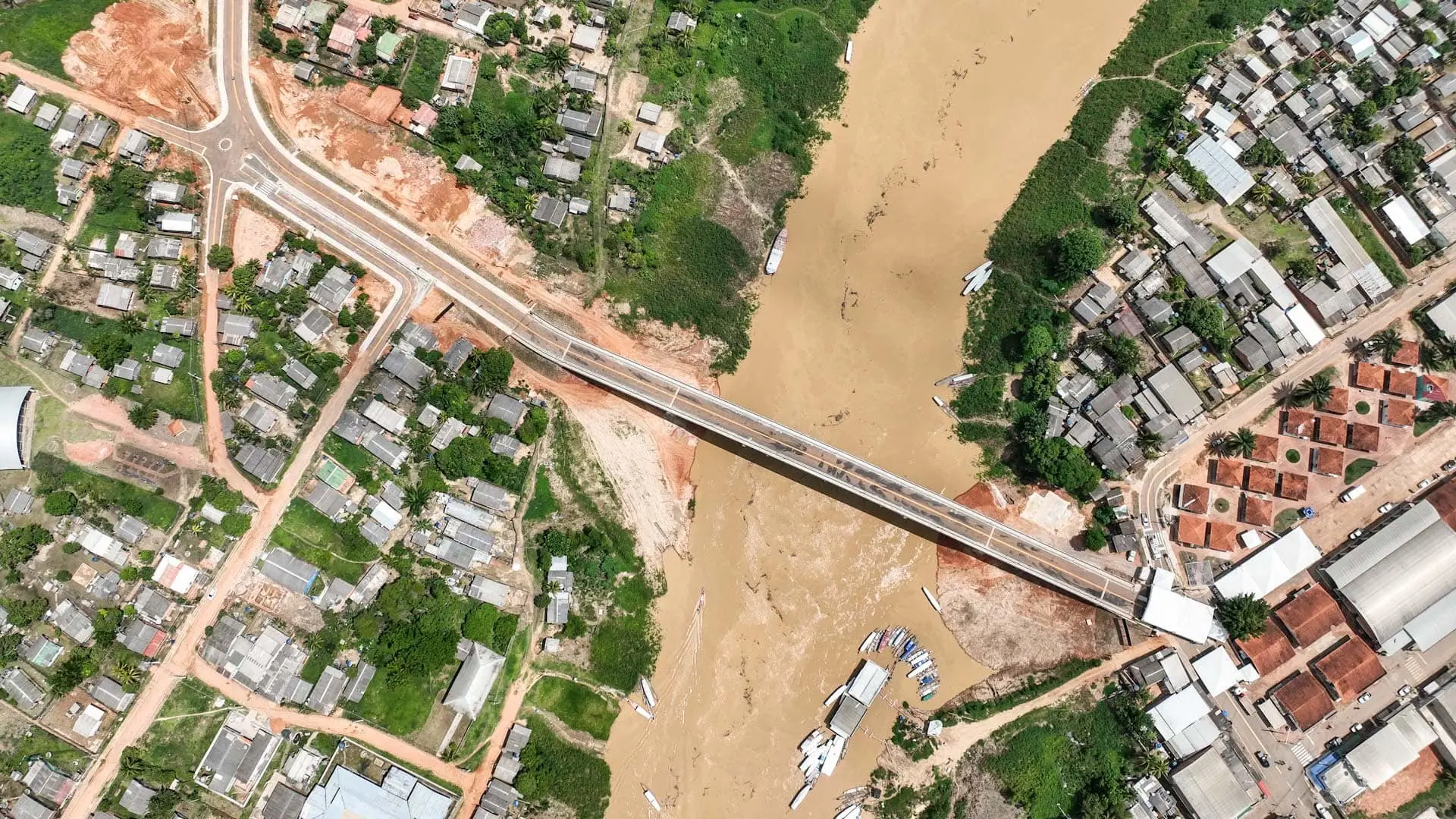 Vista aérea vertical de uma ponte de concreto recém-construída que cruza um rio de águas barrentas e marrons. A ponte conecta duas áreas residenciais urbanas com casas simples e pequenas. Na margem esquerda do rio, há uma nova estrada asfaltada com faixas brancas pintadas que dá acesso à ponte, cercada por terra batida avermelhada e vegetação rasteira. Na margem direita, casas estão dispostas de forma mais densa e há pequenos barcos atracados na beira da água. O rio corre diagonalmente pelo centro da imagem.