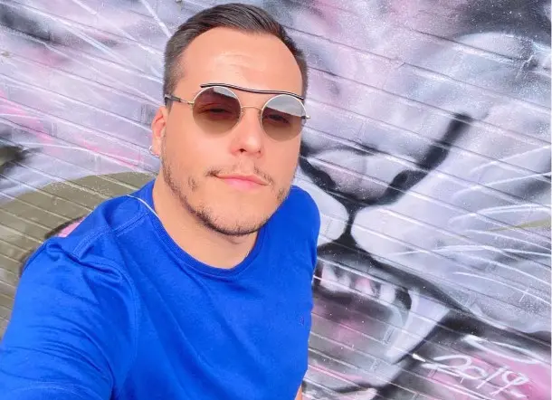 Selfie em primeiro plano de um homem de pele clara, cabelos escuros e barba curta. Ele está ligeiramente inclinado para a frente, sorrindo de forma sutil. Veste uma camiseta azul-bicicleta e usa óculos de sol redondos com lentes degradê marrom e uma armação fina dourada com uma barra preta estilizada acima das lentes. No detalhe, tem um brinco de argola pequena na orelha direita. O fundo é uma parede de tijolos pintada com um grafite expressionista em tons de cinza, preto e rosa, retratando a face estilizada de um felino de dentes afiados.