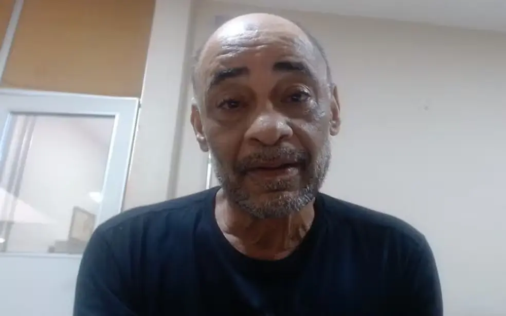 Cleomar Borges Gomes, homem de meia-idade aparece em um vídeo ou chamada virtual, olhando diretamente para a câmera. Ele veste camiseta preta e está em um ambiente interno com paredes claras e uma divisória de vidro ao fundo. A imagem é um enquadramento próximo do rosto e da parte superior do tronco.