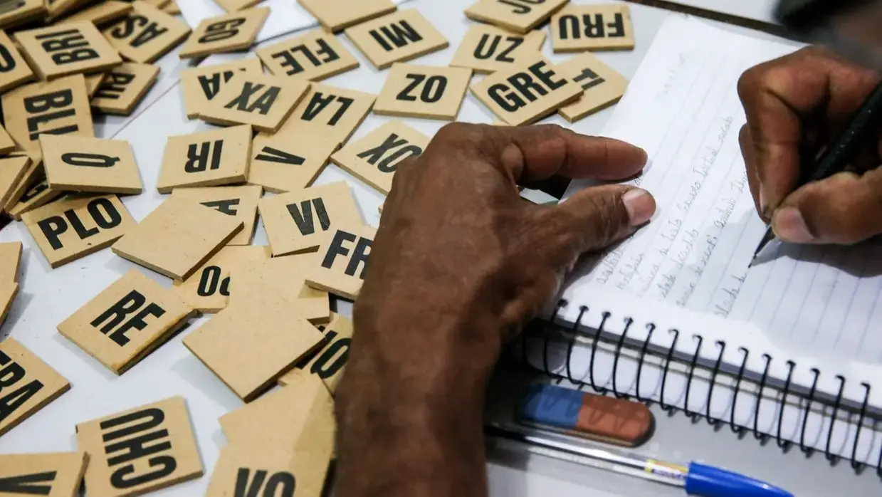 Mão segurando caneta e preenchendo formulário enquanto palavras recortadas em papel com números e letras aparecem sobre a mesa, sugerindo organização ou estudo.