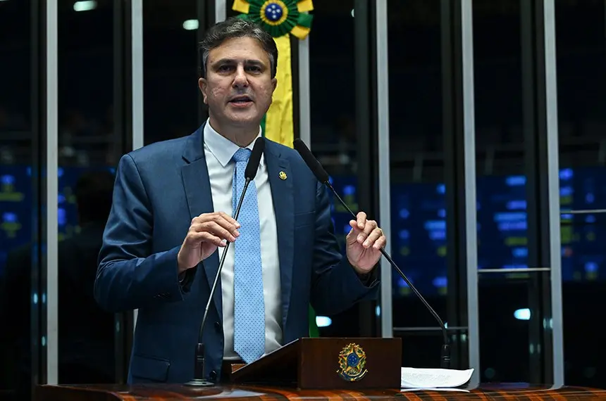Em pé na tribuna do Senado Federal, o senador Camilo Santana (PT) faz um pronunciamento durante sessão legislativa. Ele usa terno azul-escuro, camisa branca e gravata azul-clara, e gesticula com as mãos enquanto fala aos microfones. Atrás dele, a bandeira do Brasil está posicionada entre as estruturas do plenário, onde também são visíveis painéis eletrônicos iluminados.