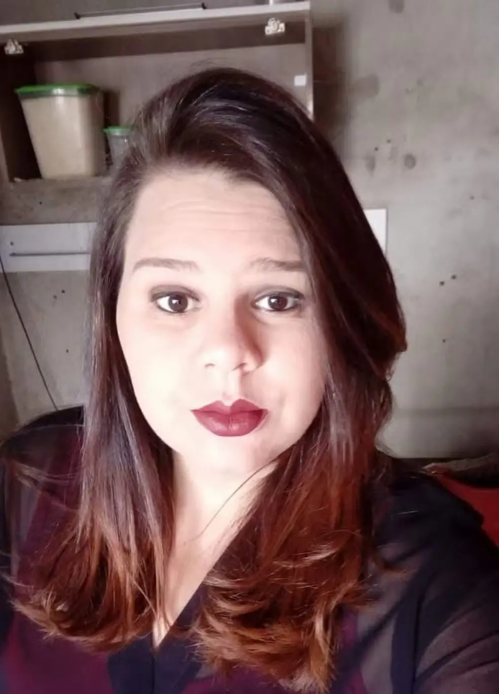 Foto em ambiente interno com mulher de cabelo castanho longo e uniforme escuro, usando maquiagem com batom vinho, encarando a câmera e com expressão surpresa. Fundo com armários.