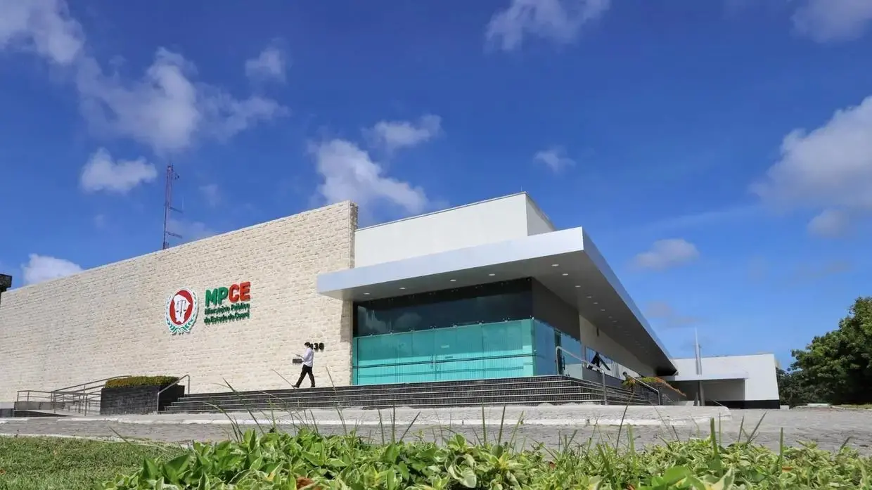 Fachada moderna do prédio do MPCE (Ministério Público do Ceará), com arquitetura contemporânea, vidro e parede clara, sob céu azul em Fortaleza.