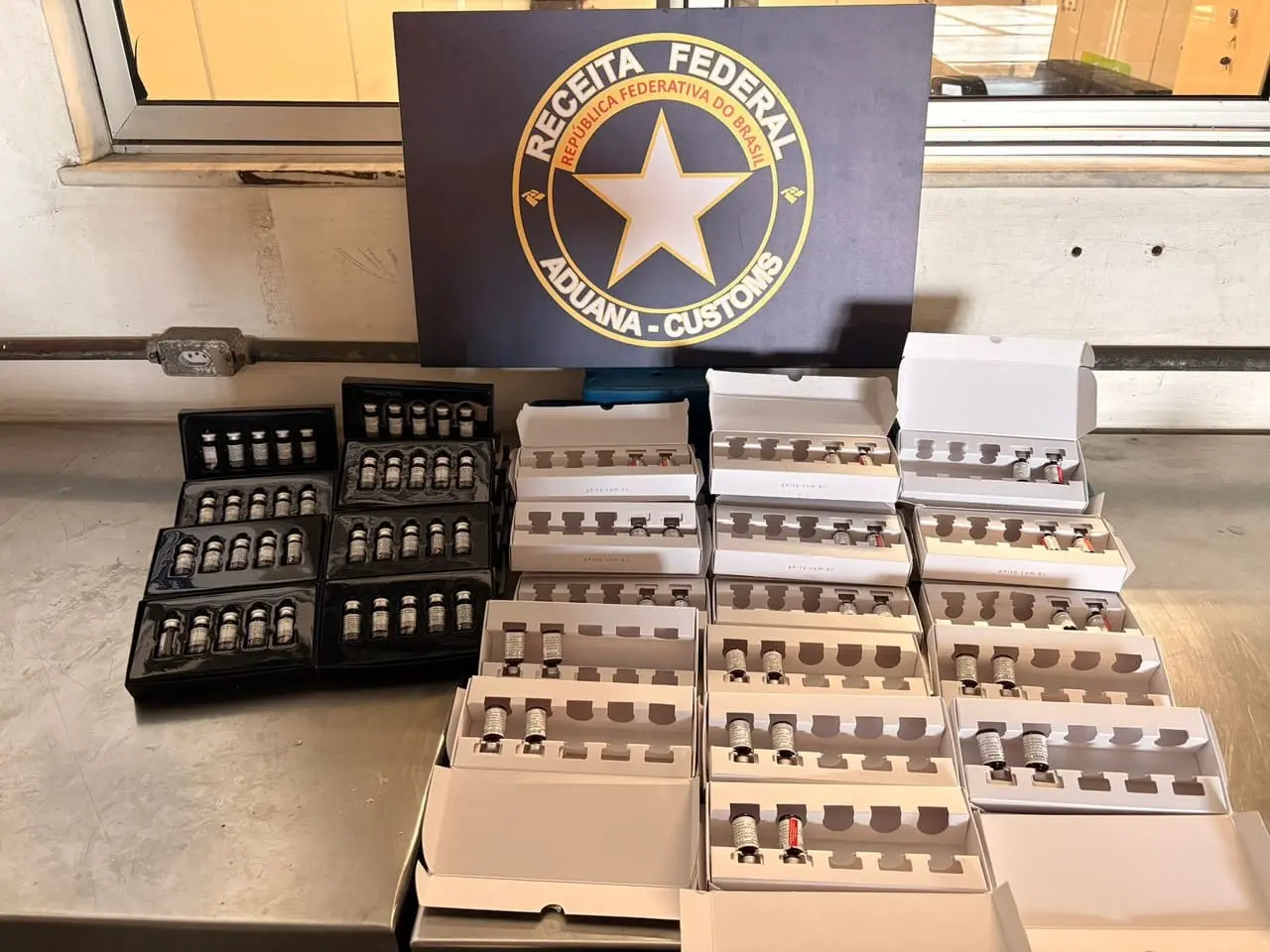 Apreensão da Receita Federal mostra dezenas de frascos de Mounjaro pequenos, organizados em caixas brancas e estojos pretos sobre uma bancada metálica, com placa ao fundo escrita Receita Federal – Aduana Customs.