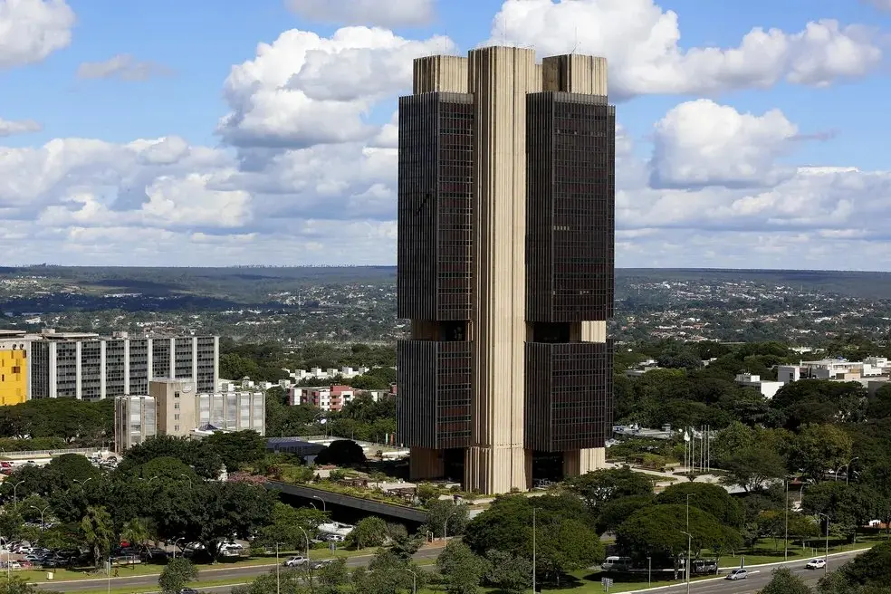 Torre de edifício alto com fachada em vidro e concreto no centro de uma cidade, com vista panorâmica de ruas, árvores e construções ao redor sob céu parcialmente nublado