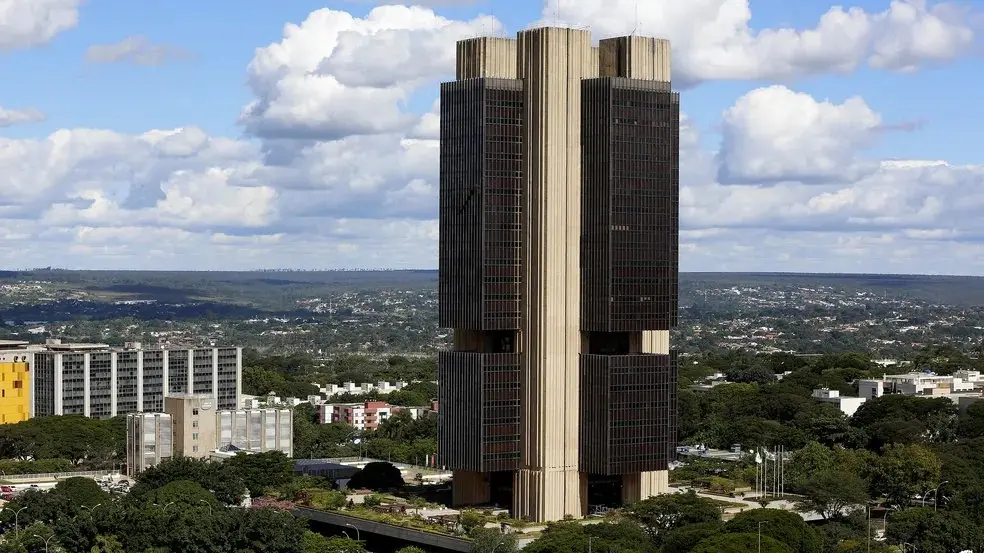 Torre de edifício alto com fachada em vidro e concreto no centro de uma cidade, com vista panorâmica de ruas, árvores e construções ao redor sob céu parcialmente nublado
