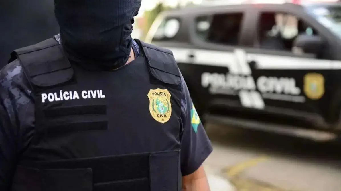 Policial civil com balaclava preta em primeiro plano e viatura da Polícia Civil ao fundo.
