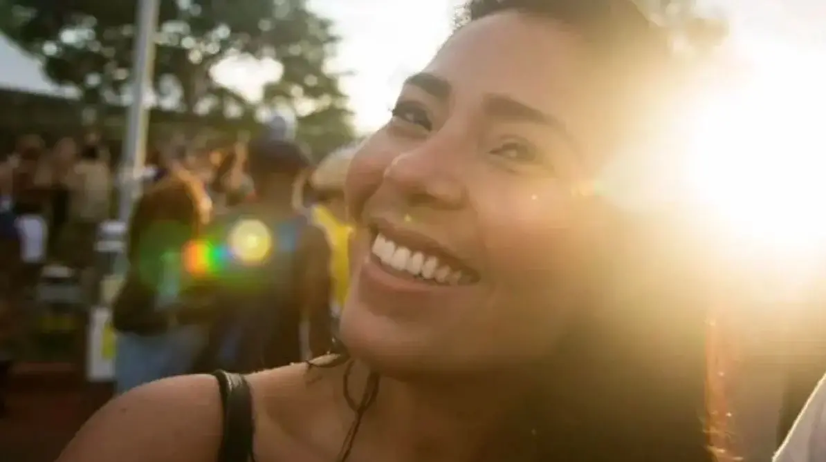 Pessoa em primeiro plano, ao ar livre, com luz do sol forte incidindo lateralmente e criando reflexos na lente. Ao fundo, há um grupo de pessoas desfocadas em um espaço aberto, com árvores e estrutura metálica visíveis.