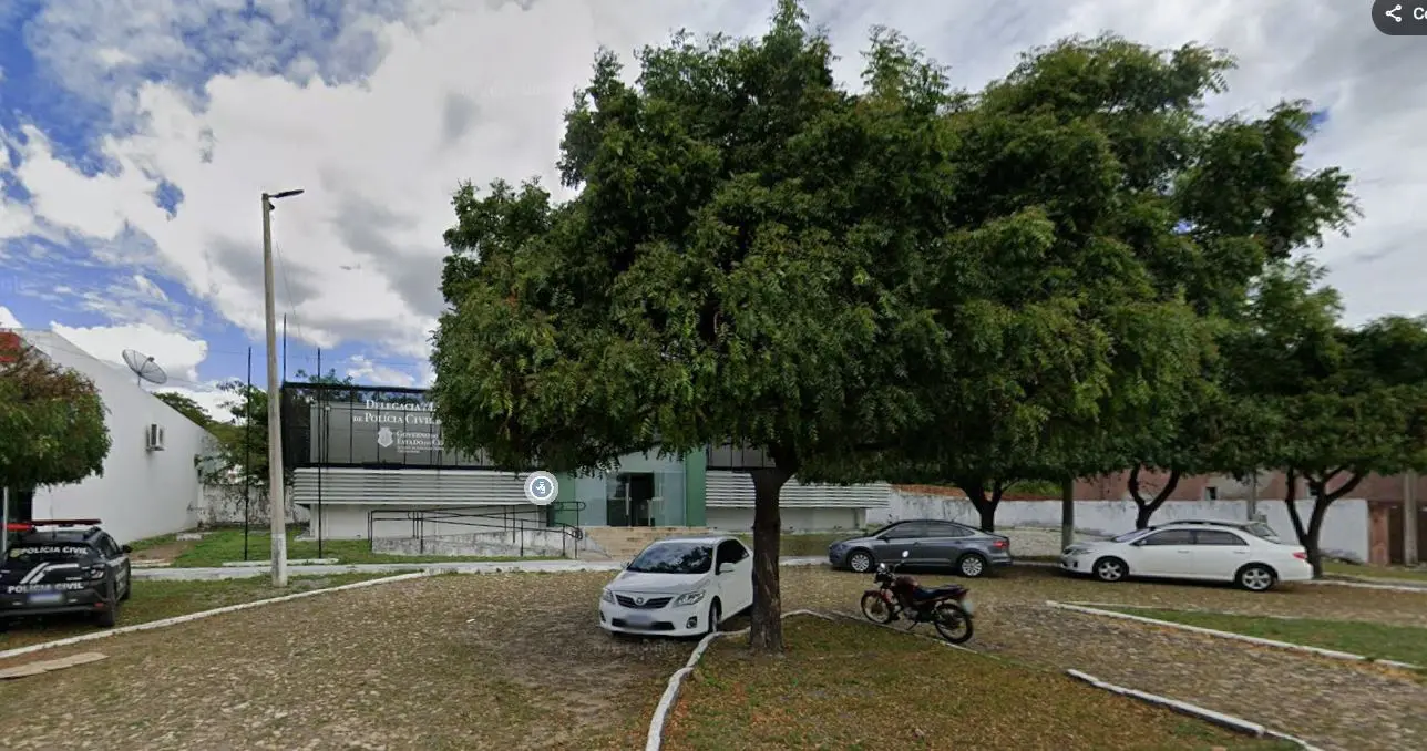 Fachada de um prédio da Polícia Civil com placa de identificação, parcialmente encoberta por árvores frondosas na frente. Em primeiro plano, há uma área pavimentada com pedras irregulares, onde estão estacionados um carro branco, um carro cinza, um carro branco maior e uma motocicleta. À esquerda, há uma viatura policial. O céu aparece com nuvens e trechos de céu azul, e um poste de iluminação está próximo ao edifício.