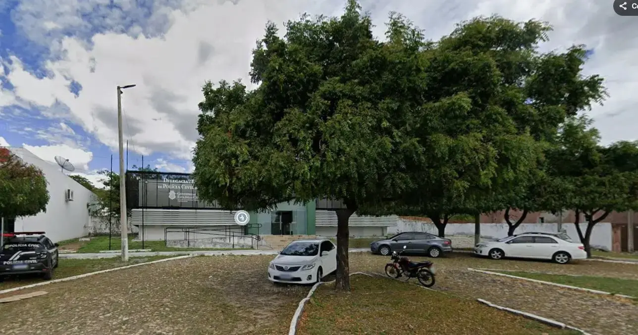 Fachada de um prédio da Polícia Civil com placa de identificação em Ipueras, parcialmente encoberta por árvores frondosas na frente. Em primeiro plano, há uma área pavimentada com pedras irregulares, onde estão estacionados um carro branco, um carro cinza, um carro branco maior e uma motocicleta. Imagem usada em matéria sobre homem morto pelo próprio irmão.