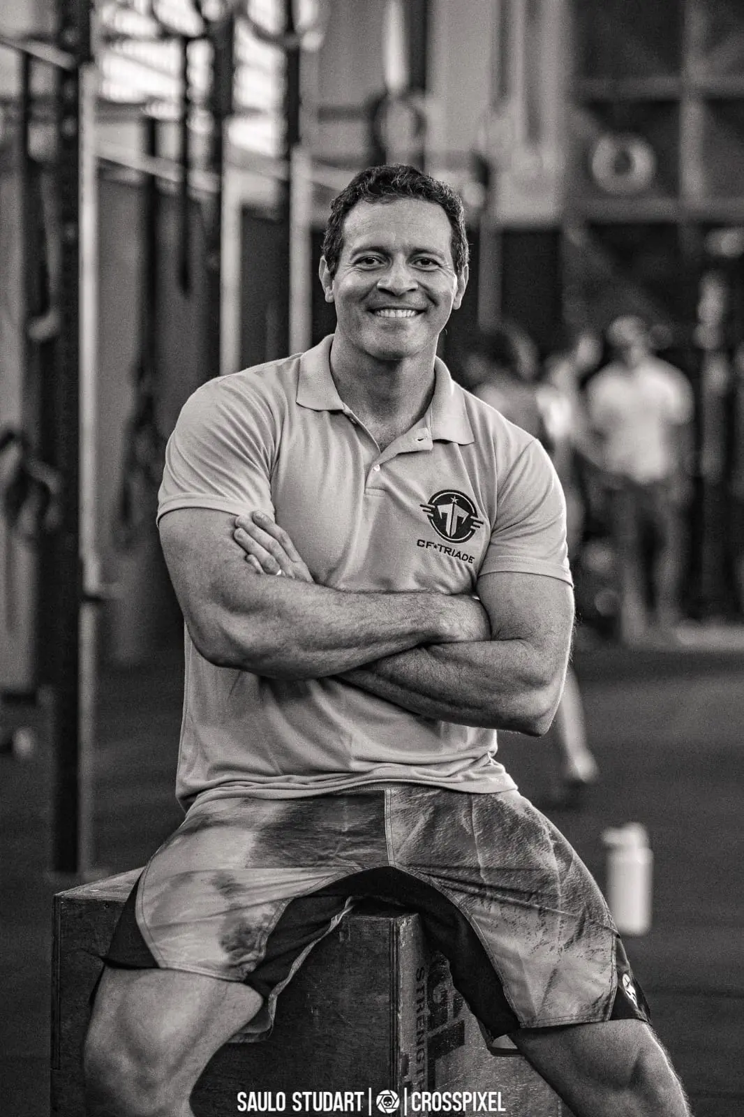 Foto em preto e branco do treinador Alberone Júnior sorrindo para a câmera e com uma blusa gola polo, dentro de um box de Crossfit.