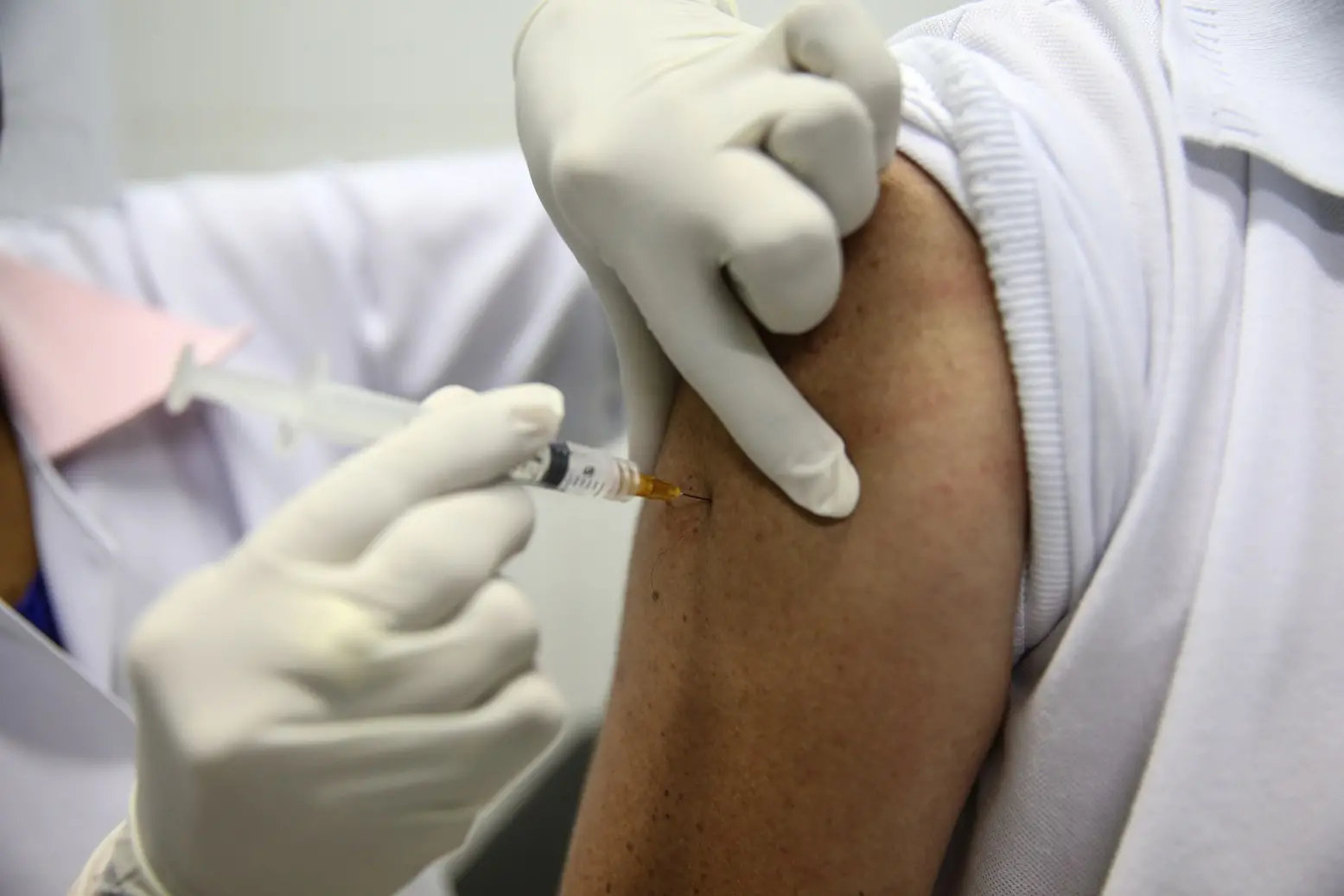 Foto em plano detalhado mostrando as mãos de um profissional de saúde, usando luvas cirúrgicas brancas, aplicando uma vacina com seringa no braço de uma pessoa. O foco principal está na agulha inserida na pele do paciente, que veste uma camisa polo branca.