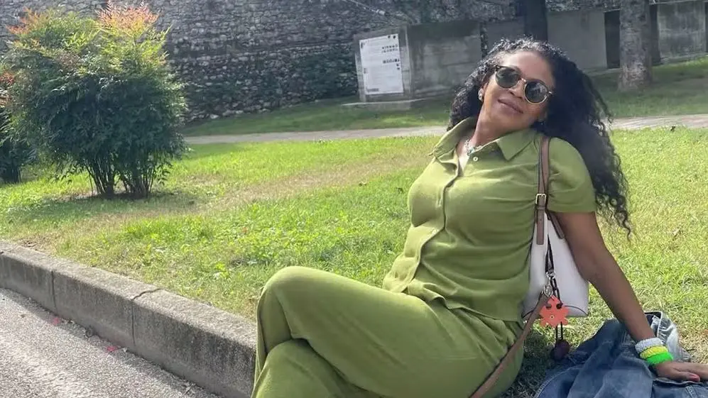 Pessoa sentada na beira de um meio-fio em área externa com gramado e árvores ao fundo. A pessoa veste conjunto verde, usa óculos escuros e segura uma bolsa de alça clara. Ao lado, há uma peça de roupa escura sobre o chão, e ao fundo aparece um muro de pedra e vegetação.