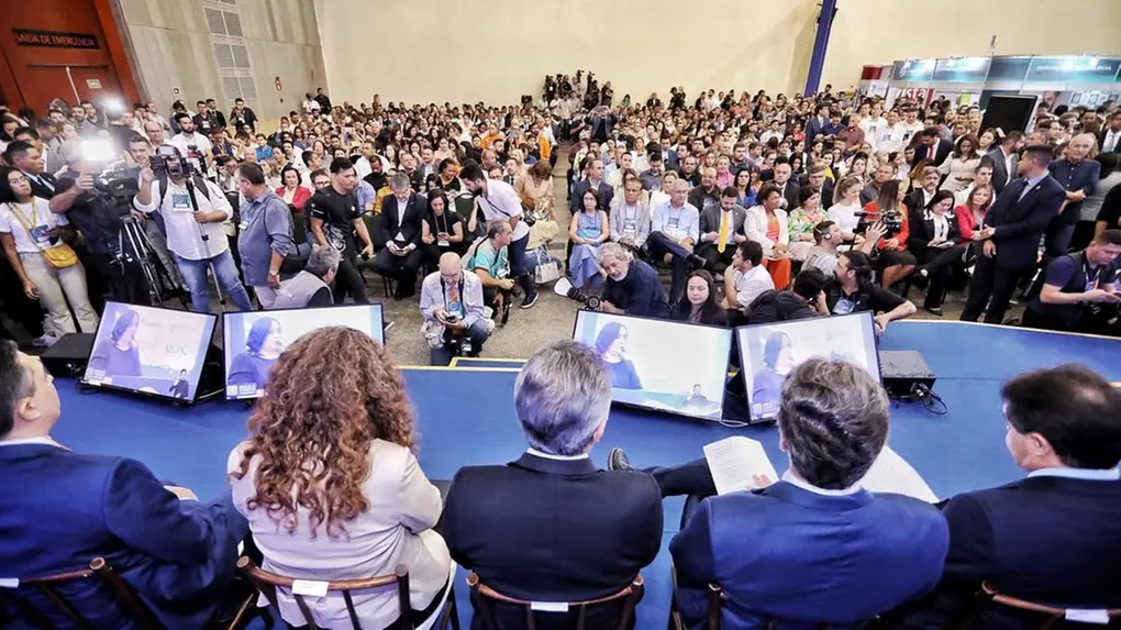 Vista ampla de auditório lotado durante evento sobre gestão pública. Em primeiro plano, autoridades e palestrantes estão sentados no palco, de costas para a câmera, enquanto centenas de participantes ocupam as cadeiras e corredores do espaço.