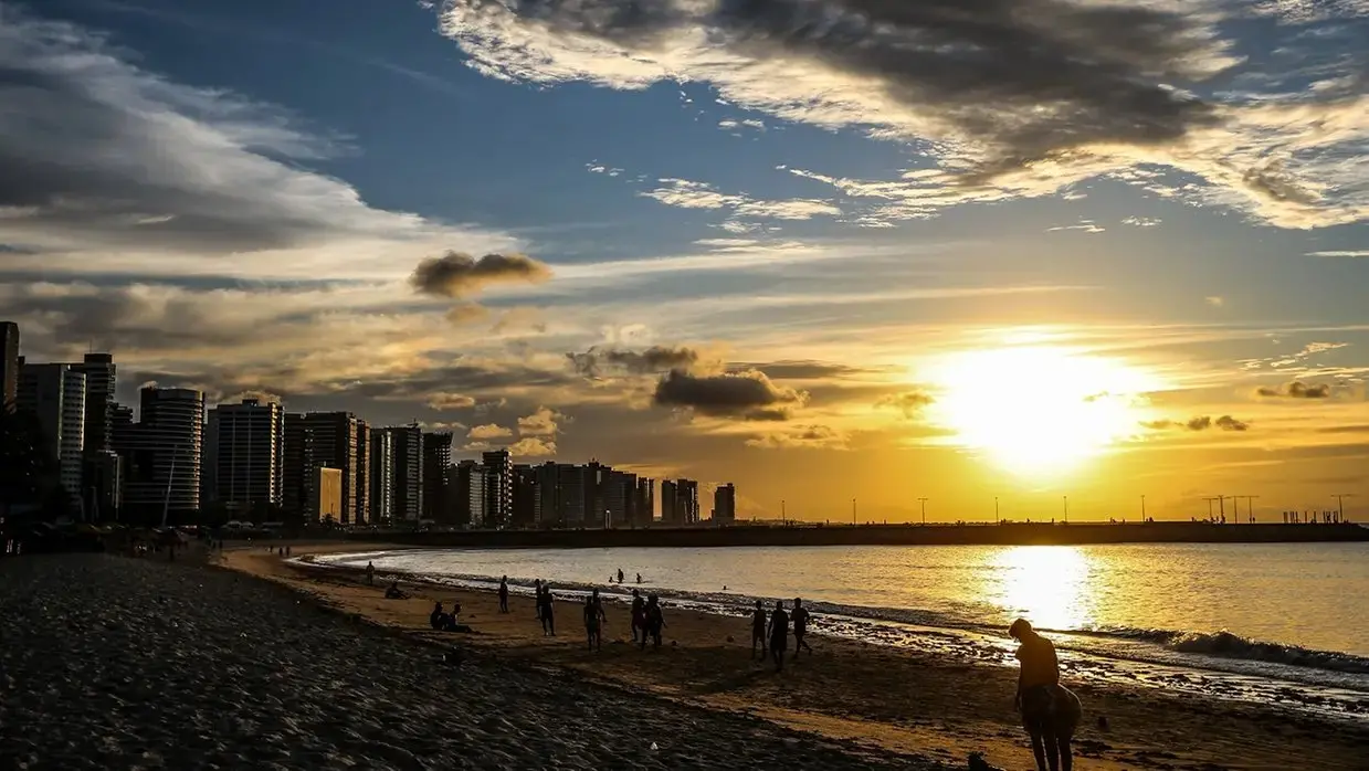 Foto tirada na orla da praia mostrando a areia, prédios ao fundo e o pôr do sol de Fortaleza.