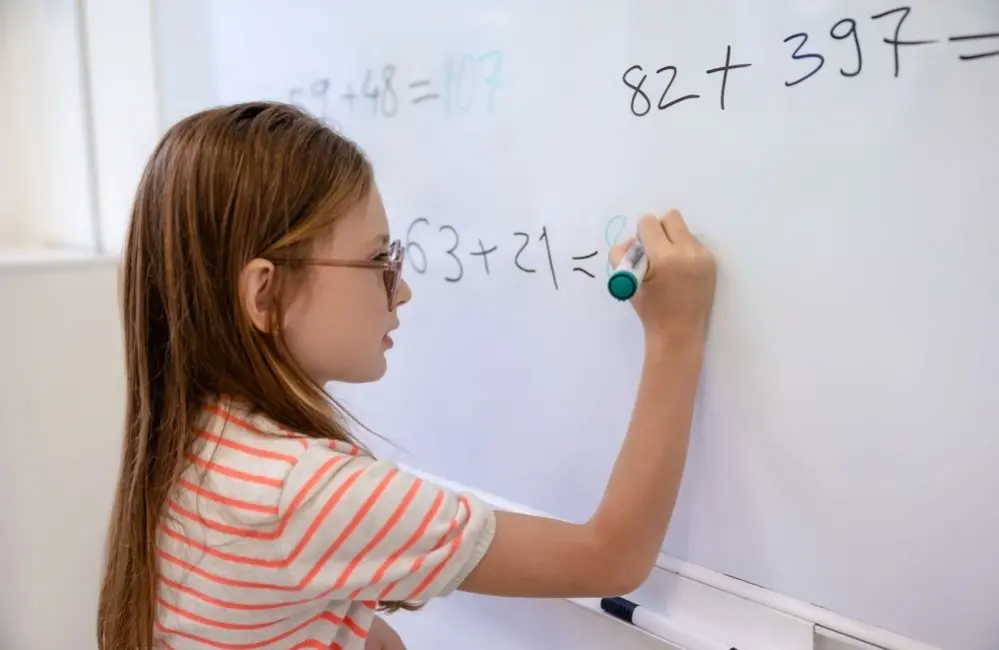 Menina faz contas de matemática em lousa branca.