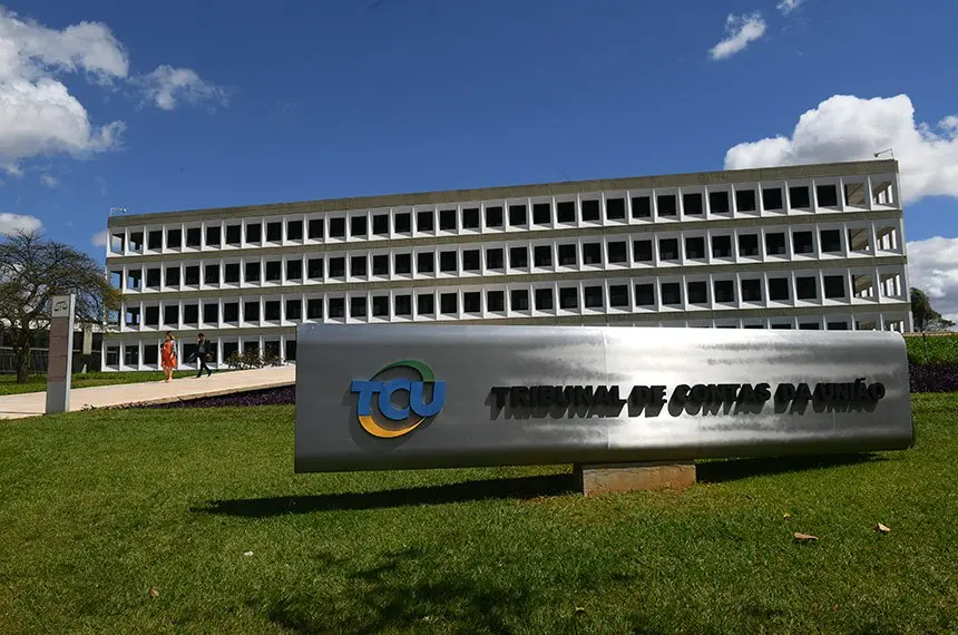 Prédio institucional branco sob céu azul com nuvens ao fundo, e em primeiro plano um letreiro do TCU — Tribunal de Contas da União sobre base de concreto em área gramada
