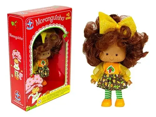 Boneca Cafezinho, da Coleção Moranguinho, em caixa e fora dela. Bonec atem o cabelo ruivo, pele morena e roupas com cores verde e amarelo predominantes.