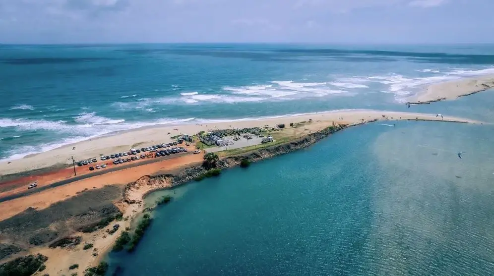 Vista aérea de uma faixa de areia entre o mar e uma lagoa, com água azul em ambos os lados. Uma estrada de terra corta a paisagem, com carros estacionados ao longo do trecho. Próximo à estrada, há estrutura com barracas e concentração de pessoas. O mar apresenta ondas suaves, enquanto a lagoa tem superfície mais calma.