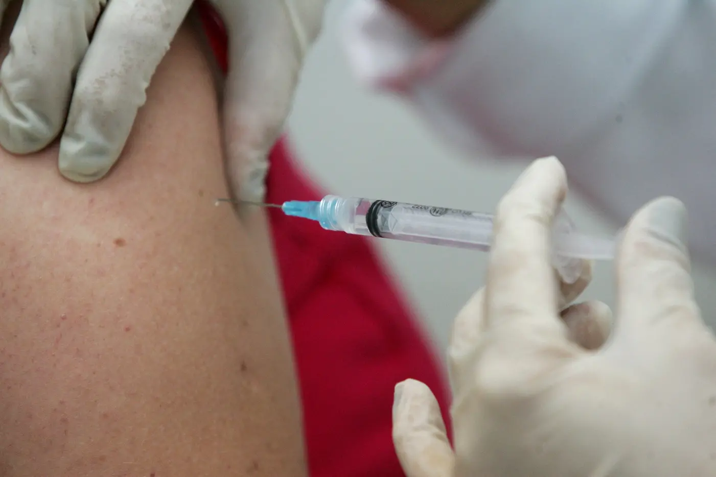 Close aproximado da aplicação de uma vacina. Mãos com luvas de plástico inserem a agulha transparente em um braço.