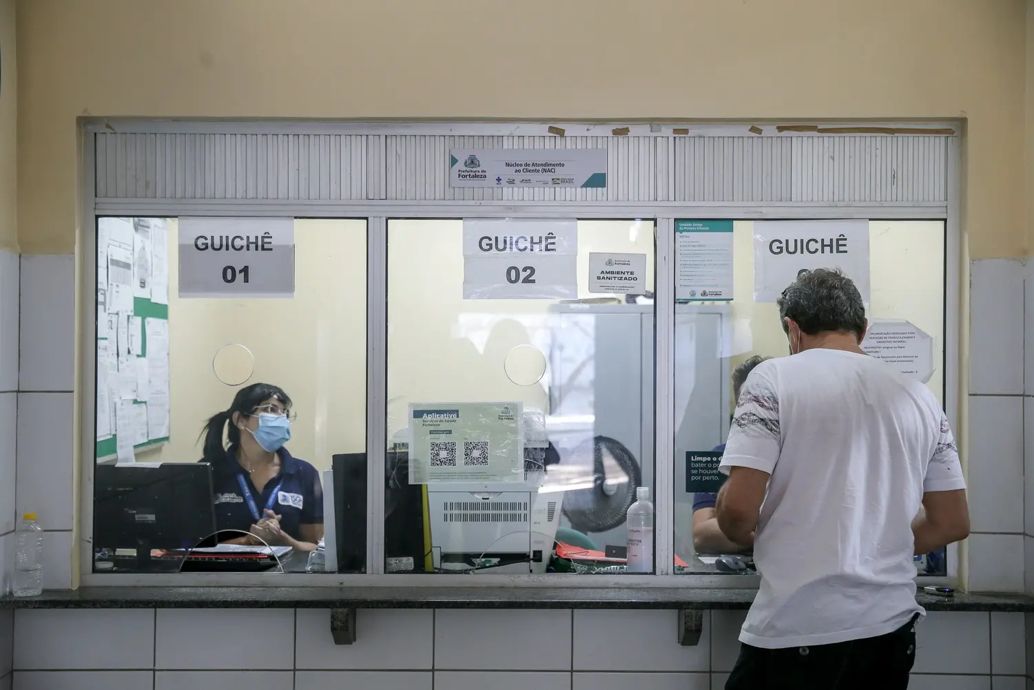 Homem de costas aguarda atendimento em frente a guichê transparente em recepção de posto de saúde da Prefeitura de Fortaleza. Dentro do guichê, há uma mulher de máscara.