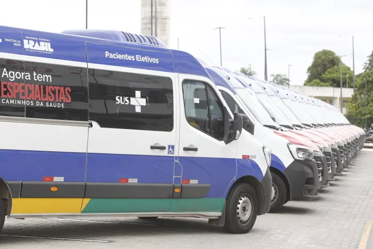 Mais de 10 vans enfileiradas em um pátio ao ar livre com adesivos do SUS e do programa Agora tem especialistas Caminhos da Saúde. Os veículos têm adesivos em cores azul, verde e amarelo.