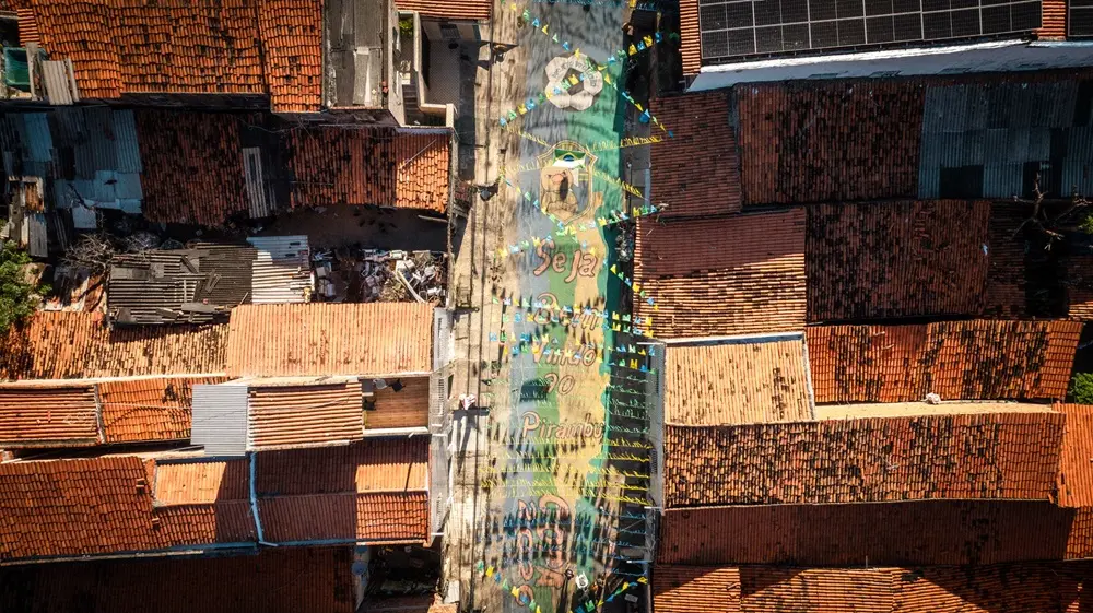 Na imagem, vista aérea vertical de uma rua estreita em uma comunidade densamente povoada. O chão da rua está pintado com o as frases: 