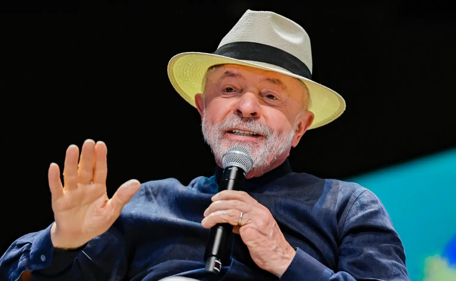 Lula, um homem idoso, de barba e cabelo brancos, usa chapéu panamá e camisa de botão azul. Ele discursa segurando um microfone.