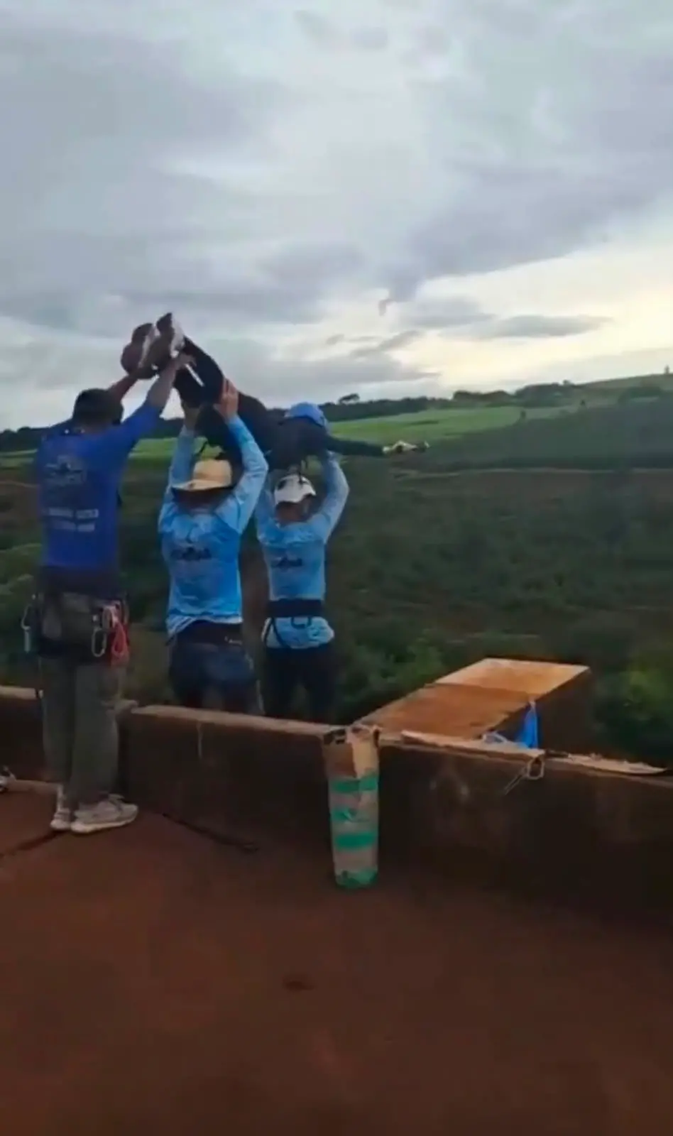 Print de vídeo em que três homens erguem uma mulher de capacete sobre os braços para jogá-la de uma ponte em atividade radical.
