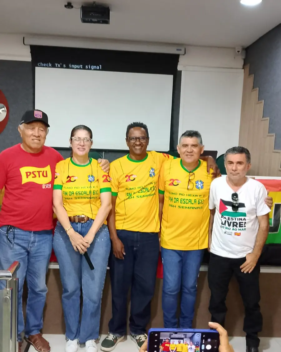 Cinco pessoas do PSTU posam para foto em evento de pré-candidatura num auditório. Um veste camisa vermelha, uma mulher e dois homens camisa verde e amarela e outro uma blusa branca com o escrito Palestina Livre.