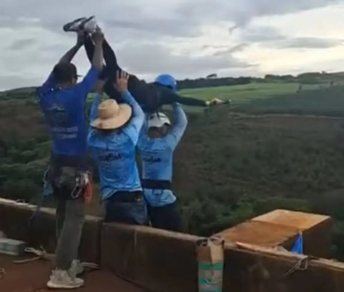 Três pessoas em área rural elevam outra pessoa pelos braços e pernas sobre uma estrutura elevada, com lavoura ao fundo e céu nublado. As pessoas usam roupas de trabalho e chapéus, e a cena acontece ao ar livre, próxima a um campo agrícola organizado em fileiras.