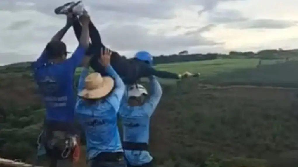 Três instrutores em área rural elevam mulher sem corda pelos braços e pernas sobre uma estrutura elevada de rope jump, com lavoura ao fundo e céu nublado. As pessoas usam roupas de trabalho e chapéus, e a cena acontece ao ar livre, próxima a um campo agrícola organizado em fileiras.