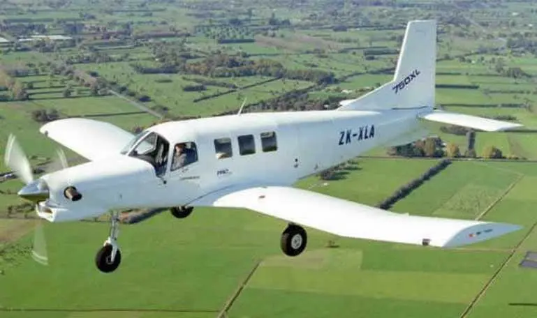 Avião monomotor modelo Pacific Aerospace 750XL branco sobrevoando campo verde.