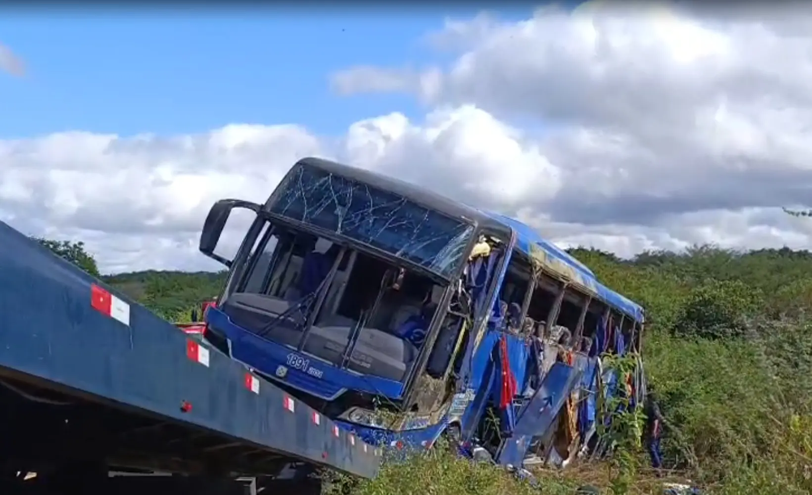 Imagem de ônibus de cor azul que tombou em Tauá, no Ceará, sendo içado por caminhão de resgate numa paisagem verde e céu nublado. O veículo tem janelas quebradas e lençóis pendendo delas.