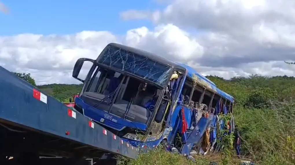 Imagem de ônibus de cor azul que tombou em Tauá, no Ceará, sendo içado por caminhão de resgate numa paisagem verde e céu nublado. O veículo tem janelas quebradas e lençóis pendendo delas.