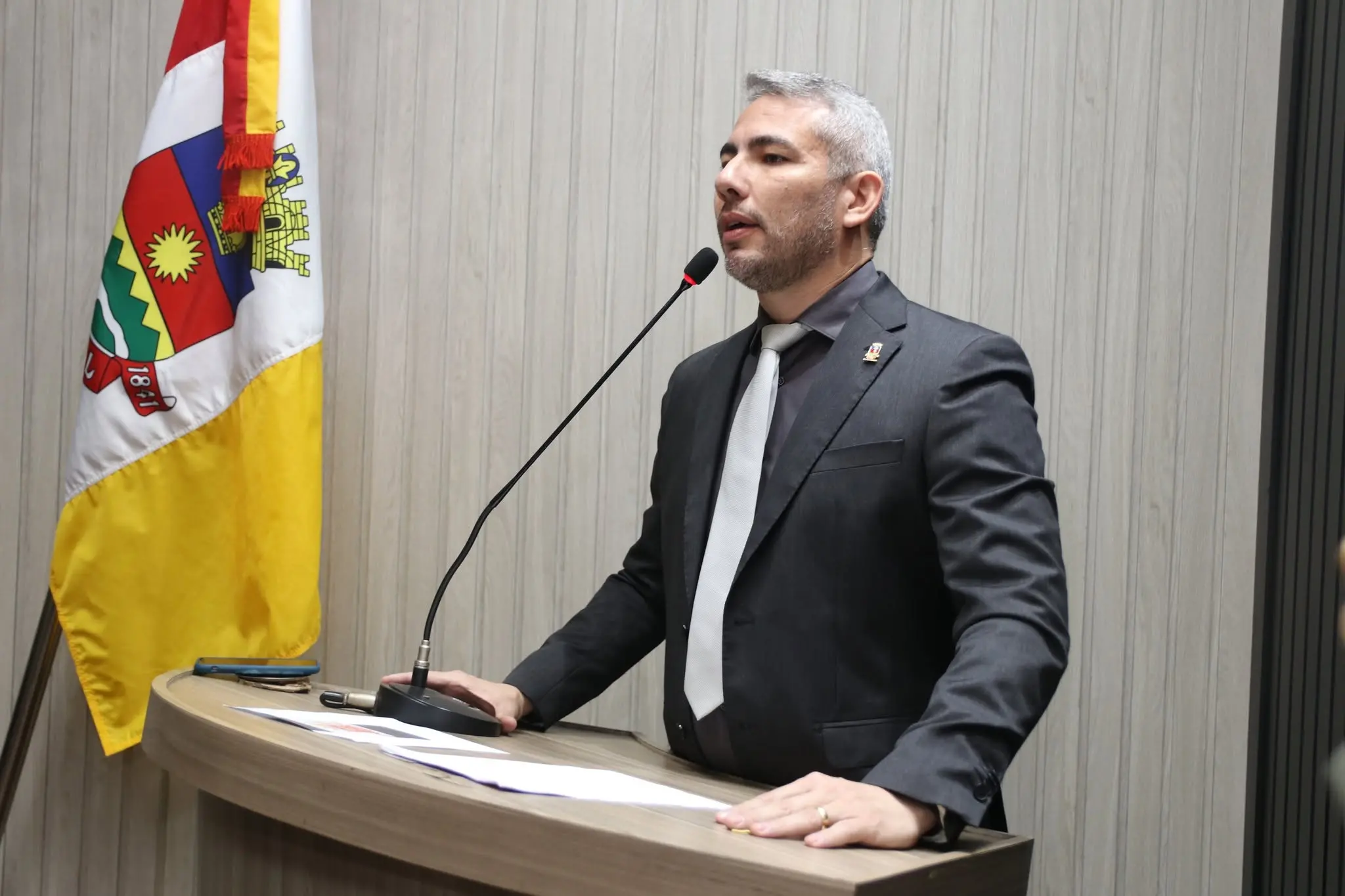Homem de cabelos grisalhos curtos e barba aparada fala ao microfone em uma tribuna de madeira, durante uma sessão em ambiente institucional. Ele veste terno escuro, camisa preta e gravata cinza clara, apoiando as duas mãos sobre o púlpito, onde há folhas de papel e um celular. Ao fundo, há uma parede com revestimento amadeirado claro e, à esquerda da imagem, uma bandeira com detalhes em vermelho, amarelo, branco, verde e azul.