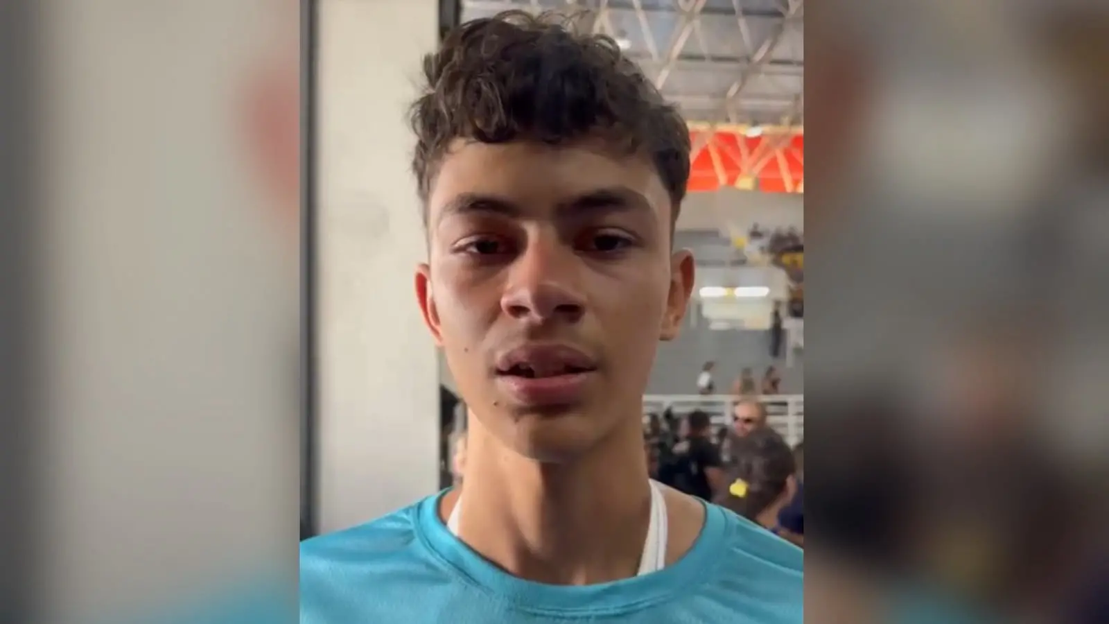 Print de vídeo mostrando o rosto de Lucas Lira, um jovem de 18 anos, pardo, magro, sem barba, de cabelos ondulados e vestindo uma camisa azul. Ao fundo, há um ginásio poliesportivo.