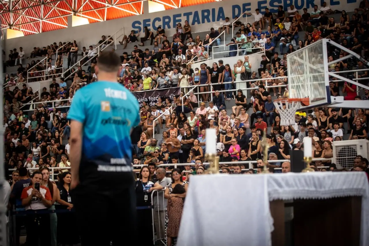 A imagem mostra um homem em primeiro plano de costas, de camisa azul onde está escrito técnico. À direita, uma mesa coberta com um pano branco e algumas velas acesas. O homem e a mesa parecem estar em uma quadra de basquete devido a presença de uma tabela e aro de basquete. Ao fundo, uma arquibancada repleta de pessoas olhando atentamente para a direção do homem. Na parte de cima, está escrito secretaria de esporte.